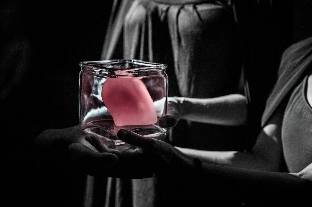 Μερικώς ασπρόμαυρη φωτογραφία που απεικονίζει πέντε χέρια που κρατάνε ένα γυάλινο κυβικό δοχείο που περιέχει ένα έντονο ροζ σφαιρικό αντικείμενο. Η εικόνα συνοδεύει το κείμενο «Χώρος».